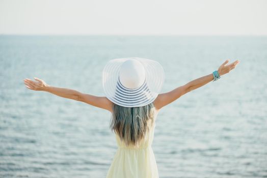 Happy young woman wearing in dress and hat with a wide brim standing with raised arms and enjoying view of sea, rear view