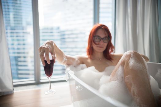 A ginger woman lying in the foam bath and hold a glass of red wine - a view on a modern glass buildings from the panoramic windows. Mid shot
