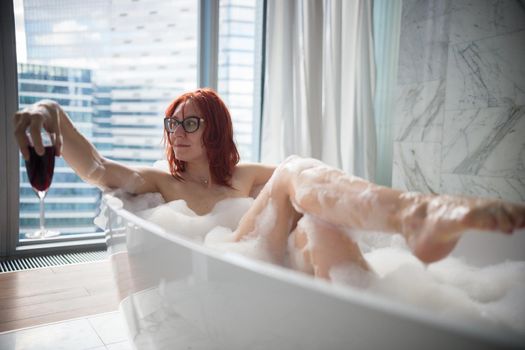 A ginger woman lying in the foam bath and looking to the side - holding a glass of red wine - a view on a modern glass buildings from the panoramic windows. Mid shot