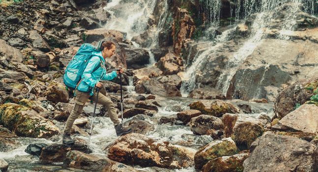 Backpacker young woman crossing mountain river with trekking poles near the waterfall