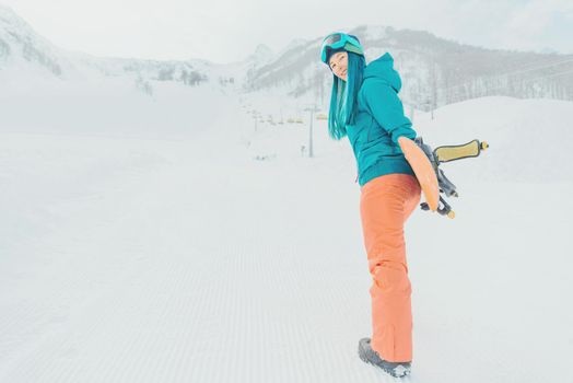 Beautiful young woman with snowboard standing near slope on ski resort, looking at camera