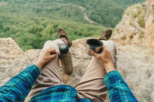 Female traveler sitting on cliff and drinking tea from thermos in the mountains, point of view
