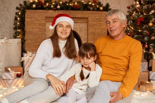 Portrait of happy family, mature man wearing orange sweater, his daughter and granddaughter sitting near Christmas tree at home, looking at camera with positive facial expressions.