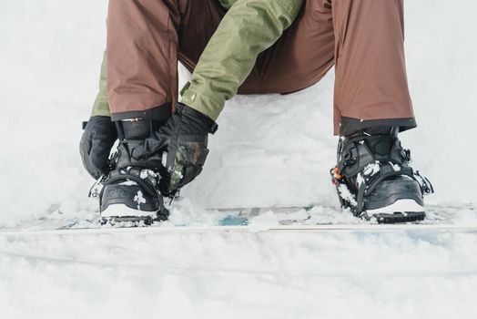 Unrecognizable young man putting on his snowboard and tightening the straps on background of snow in winter