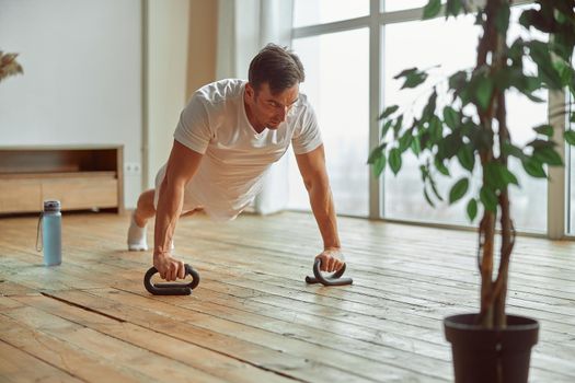 Strong male is doing workout at home and staying in plank position with professional handles