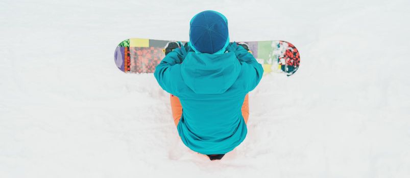 Unrecognizable young woman sitting with snowboard on snow, rear view