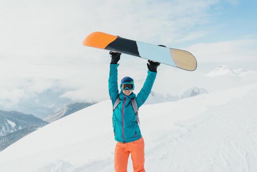 Happy young woman holding snowboard over her head on snowy mountains in winter, looking at camera