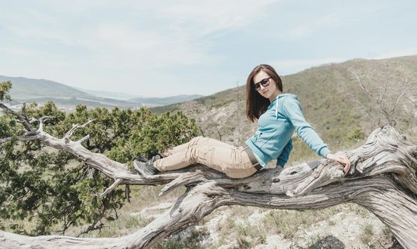 Beautiful girl in sunglasses resting on juniper tree outdoor, looking at camera