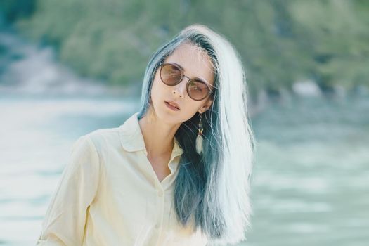 Portrait of attractive young woman with blue hair in sunglasses on background of lake, looking at camera.