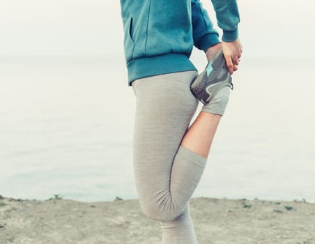 Unrecognizable sporty young woman stretching her legs and preparing to run on coast in summer.