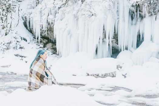 Beautiful young woman wrapped in plaid resting on the river bank in winter.