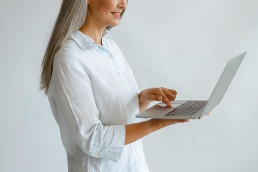 Cheerful hoary haired middle aged lady in white blouse uses modern laptop on light background in studio side view closeup