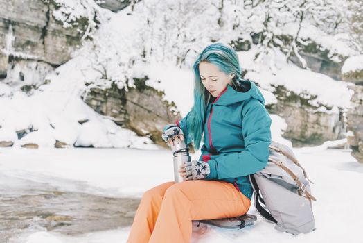 Hiker young woman resting on the river bank and opening a thermos to drink tea in winter.