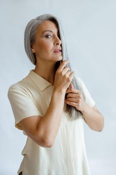 Attractive middle aged Asian model in hides half of face with silver hair standing on light background in studio. Mature beauty lifestyle