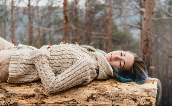 Beautiful young woman with closed eyes lying on fallen tree trunk in the woods
