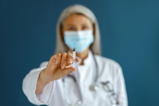 Grey haired Asian woman doctor in white coat with disposable mask stands on blue background in studio, focus on hand with syringe. Vaccination day