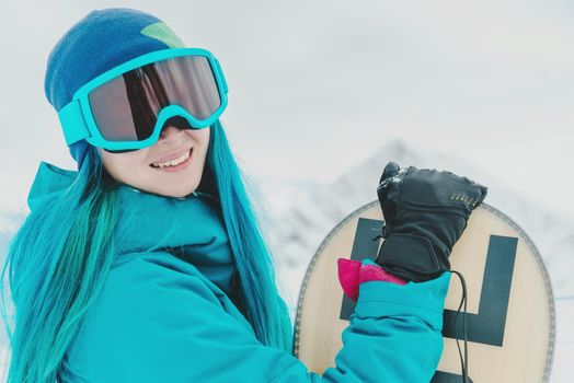 Smiling young woman with blue hair in protective sunglasses standing with snowboard on background of snowy mountains, looking at camera