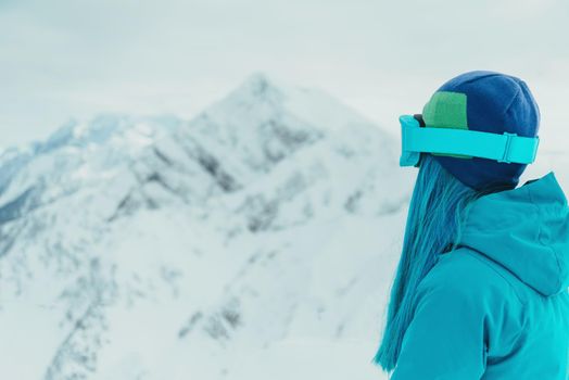 Young woman with blue hair in sunglasses looking at snowy mountains in winter