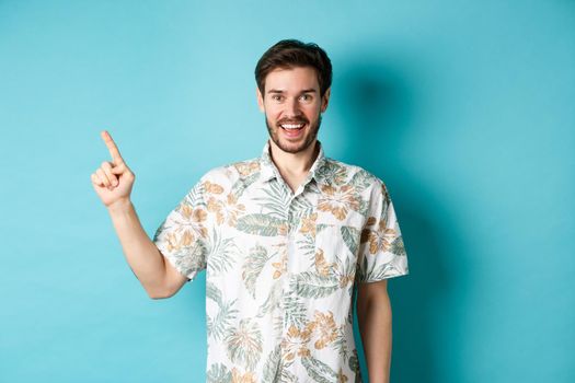 Cheerful young man in hawaiian shirt pointing finger left, showing empty space and smiling. Concept of tourism and vacation.