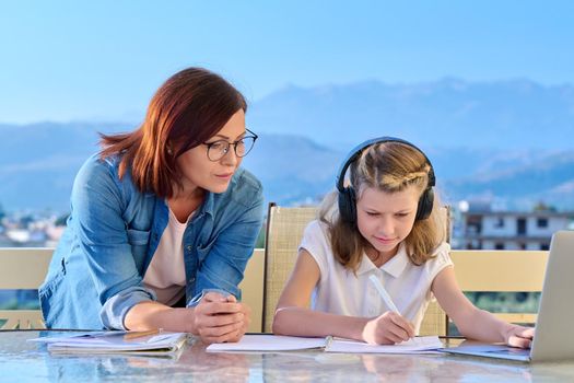 Mother helping daughter schoolgirl with online studies. Mom and girl in headphones with laptop notebooks together at home outdoor terrace. Parent child relationships, technology in school, e-learning