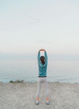 Fitness young woman stretching her hands and back on coast on background of sea, rear view