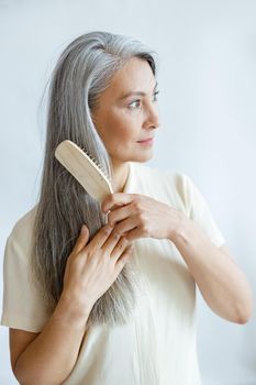 Pretty middle aged Asian person brushes straight grey hair posing on light background in studio. Mature beauty lifestyle