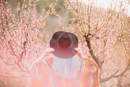 Fashionable young woman in hat walking in blossoming peach tree garden in spring, rear view.