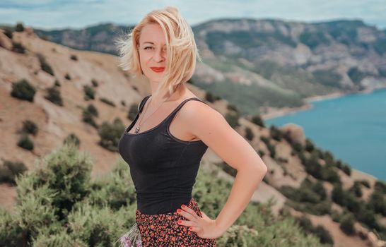 blonde girl with scarlet lipstick and red manicure in a black T-shirt with a bouquet of wildflowers on a background of sea and mountains. she is happy and enjoying wonderful breathtaking landscape.