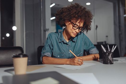 Happy Afro American lady sitting at the desk and writing on document while working in modern office
