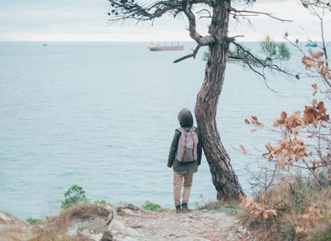 Unrecognizable female traveler with backpack standing near a tree and looking at sea, rear view. Space for text in left part of the image.