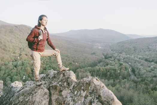 Traveler explorer girl with backpack standing on peak of cliff in the mountains and looking into the distance in summer
