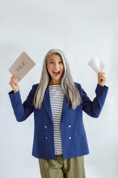 Excited mature Asian model with long hoary hair in blue jacket holds gift card and Sale sign posing on light grey background in studio