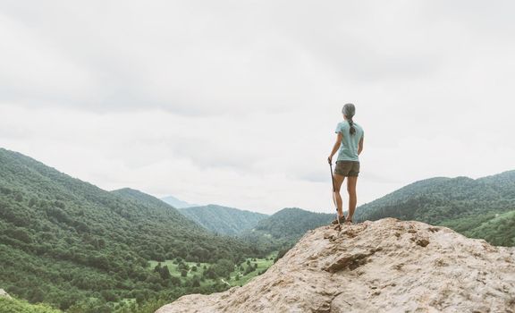 Traveler explorer young woman standing on rocky stone with trekking poles and looking at mountain valley in summer, rear view.