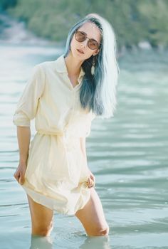 Attractive young woman with blue hair wearing in dress and sunglasses resting on lake, looking at camera.