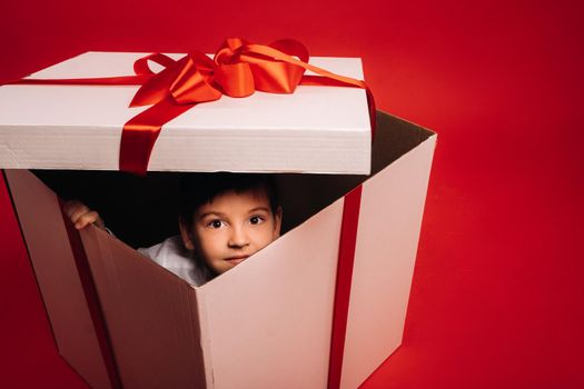 A little boy sits in a Christmas present and looks out of it on a red background.