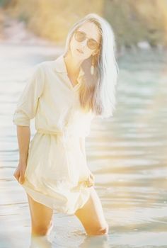 Attractive young woman with blue hair wearing in dress and sunglasses resting on lake, looking at camera. Image with sunlight effect.