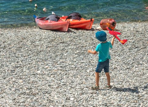 Little baby boy is playing with toy plane on the peables beach at suny day on background of kayaks. Concept of active travel by plane with children. Family holidays.