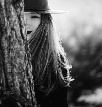 Beautiful young witch with blue hair in black dress and hat looks out from behind a tree. Monochrome image