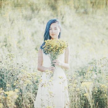 Young woman with blue hair and a bouquet of yellow flowers standing in a spring field, looking at camera. Image with old textured effect.
