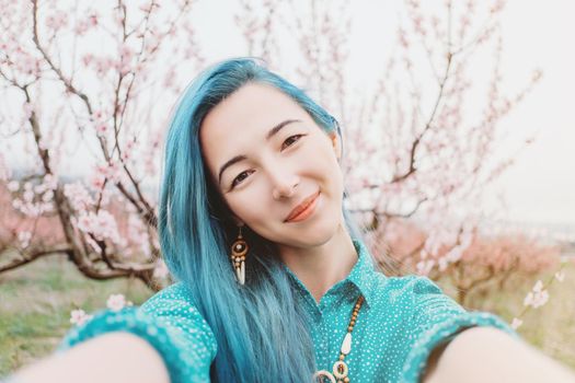 Smiling beautiful young woman with blue hair taking selfie in blossoming spring garden.
