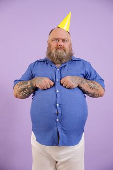 Funny displeasured mature man with overweight in tight shirt and party hat poses on purple background in studio