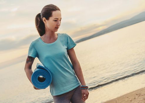 Young beautiful woman with yoga mat standing on beach in summer and ready for workout.