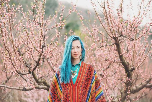 Beautiful young woman with blue hair wearing in poncho walking among the flowering pink trees in spring.