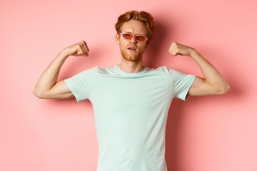 Confident young man with red hair, wearing summer sunglasses and t-shirt, showing strong and fit body muscles, flex biceps and staring cool at camera, pink background. Workout concept