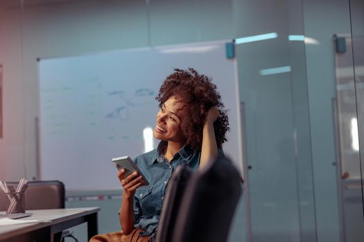Happy Afro American businesswoman sitting at workplace and holding smartphone while looking away. Copy space