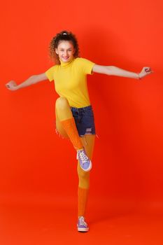 Girl in sports outfit jumping on one leg. Joyful girl with ponytail looking stylish in turtleneck blouse, leggins, shorts and socks gaiters jumping with her arms outstretched against red background