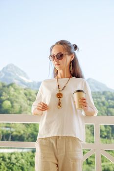 Beautiful young woman wearing in white clothing with wooden accessories resting with cup of coffee on balcony on mountains resort in summer.