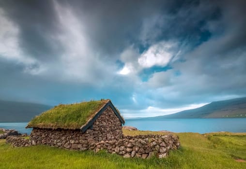 Small house near a blue lake, a small house made of stones with covered grass on the shore of a beautiful lake
