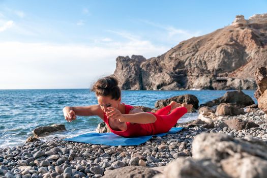 The woman in a red suit practicing yoga on stone at sunrise near the sea. Young beautiful girl in a red bathing suit sits on the seashore in lotus position. Yoga. Healthy lifestyle. Meditation