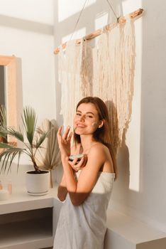 Happy young woman applying cream in bathroom. Skincare, treatment concept. Close up portrait.